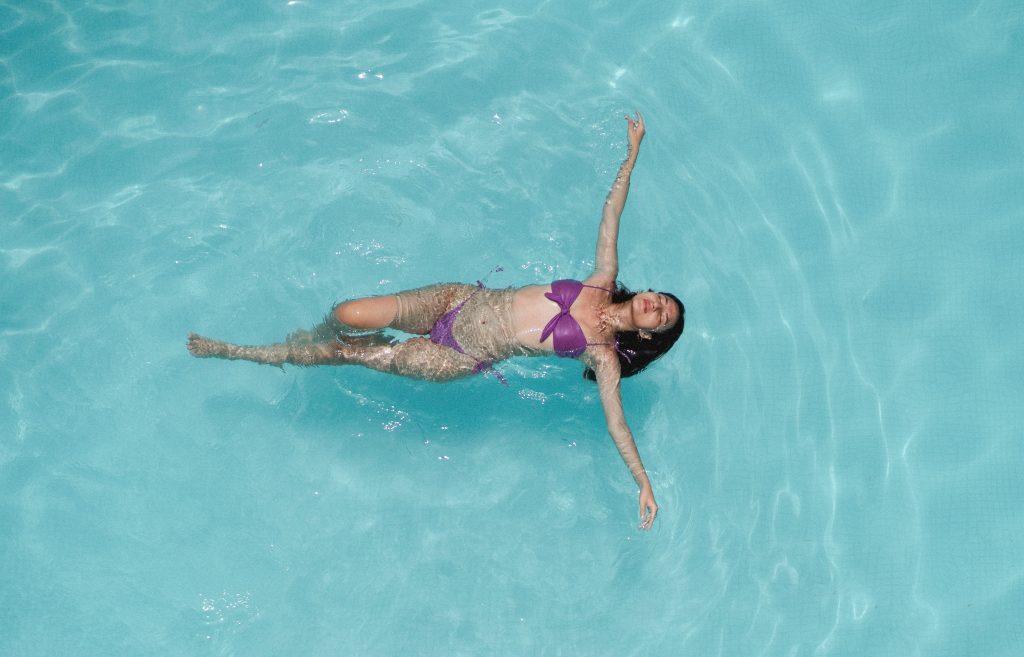 A woman in a purple bikini floats tranquilly in a clear pool, viewed from above, symbolizing relaxation and vacation vibes.